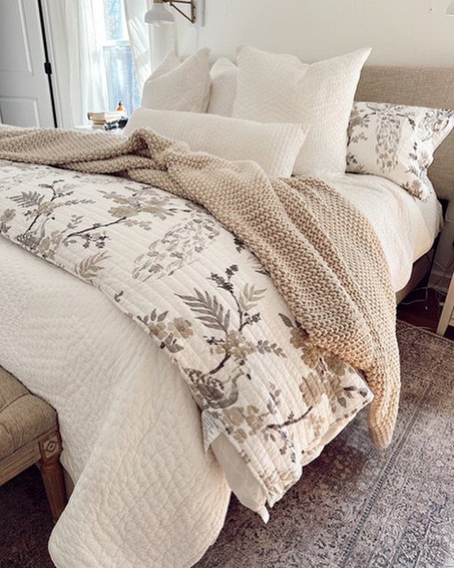 Layered bedding with floral patterns, white quilted textures, and a chunky knit throw on a bed.