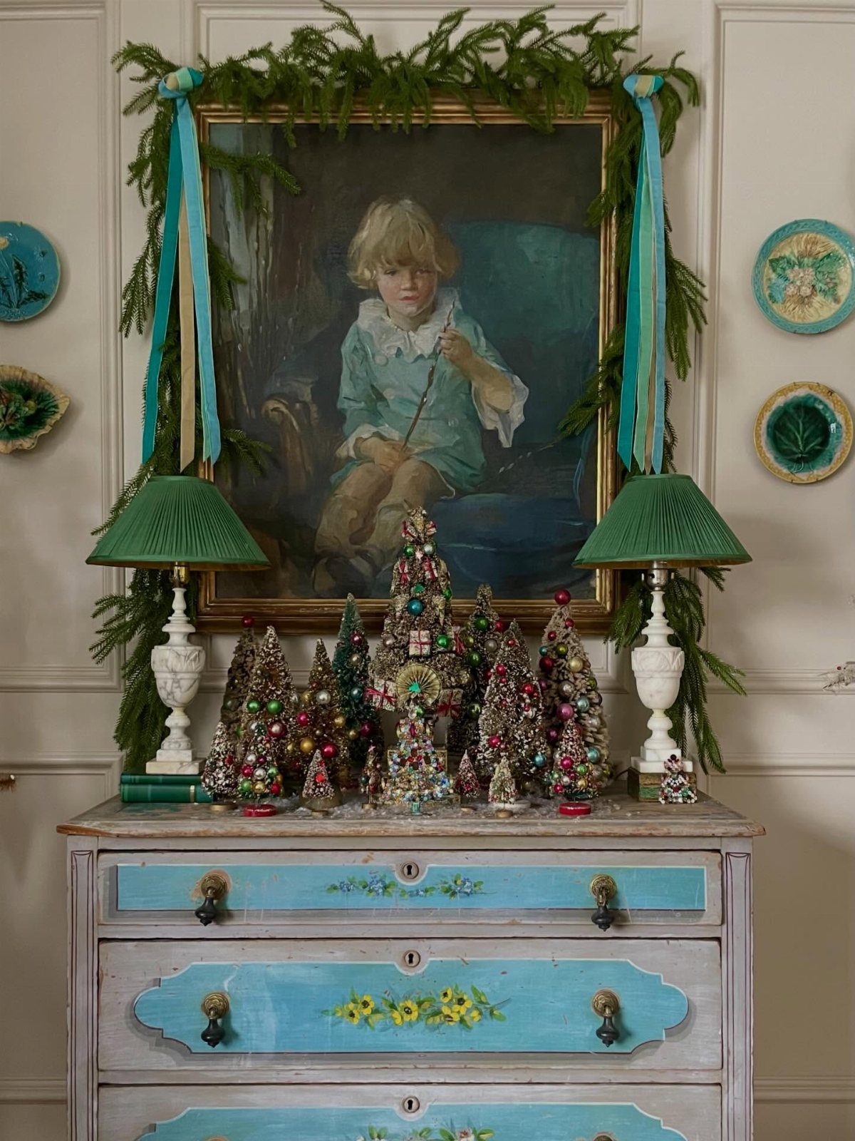 Colorful glittery bottle brush trees arranged on a vintage blue dresser under a portrait.