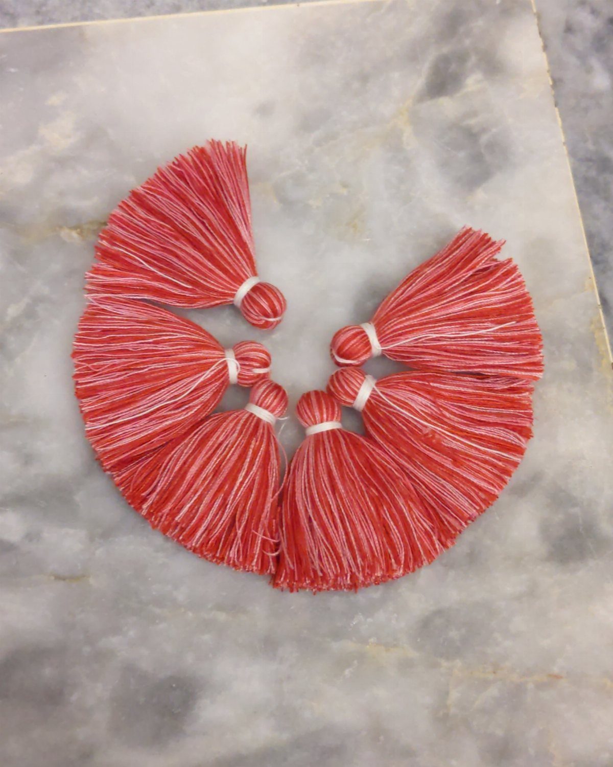 Six coral-colored handmade tassels with white thread arranged on a marble surface.