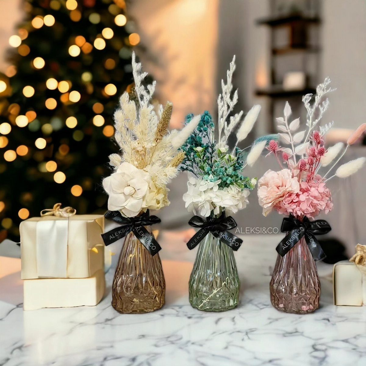 Three small glass vases with dried floral arrangements in cream, teal, and pink on marble.