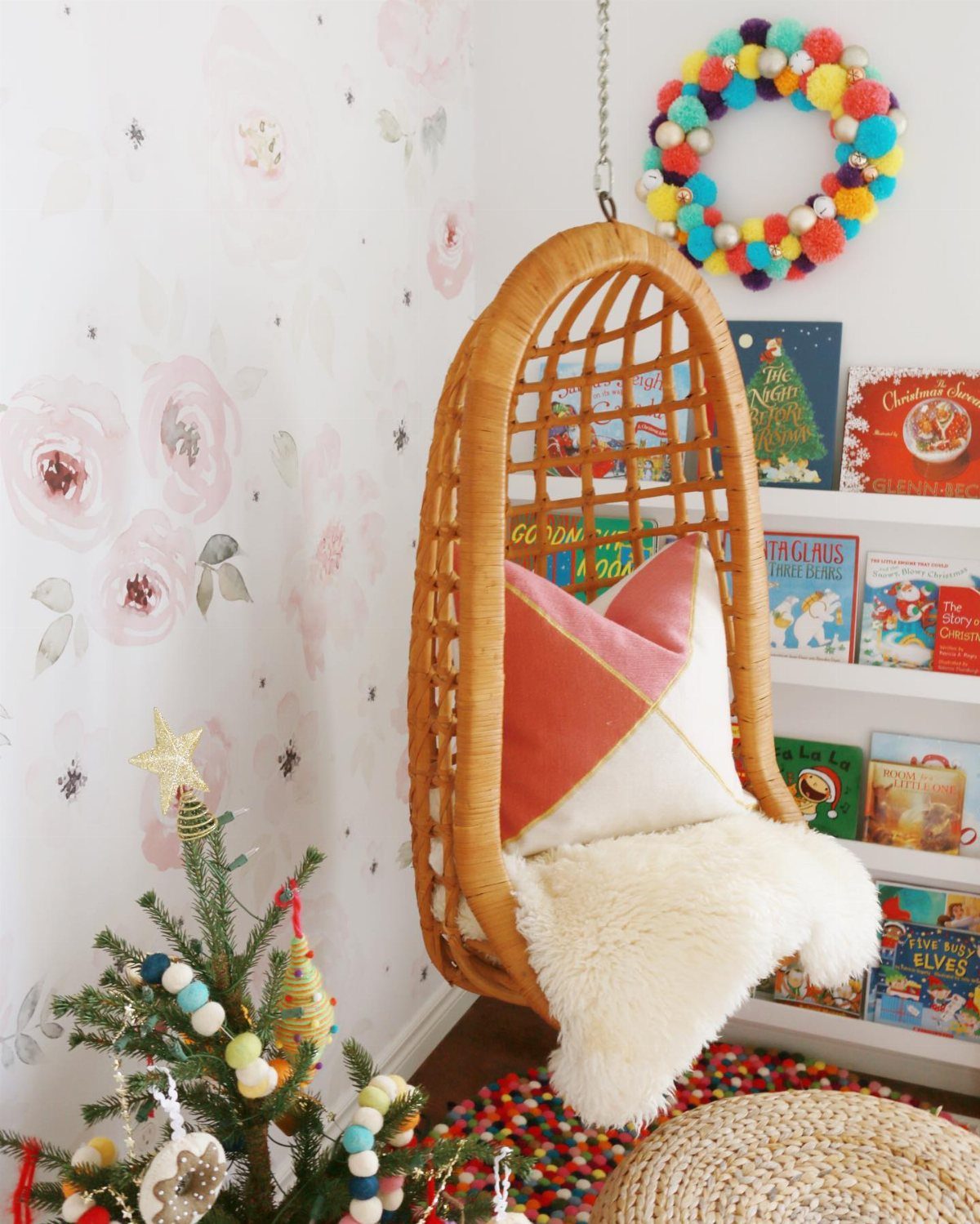 A colorful pom pom wreath hanging on a white wall above a wicker chair.