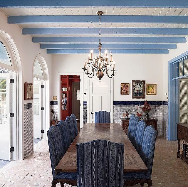 A long wooden dining table with blue upholstered chairs under a brass chandelier in a bright room.