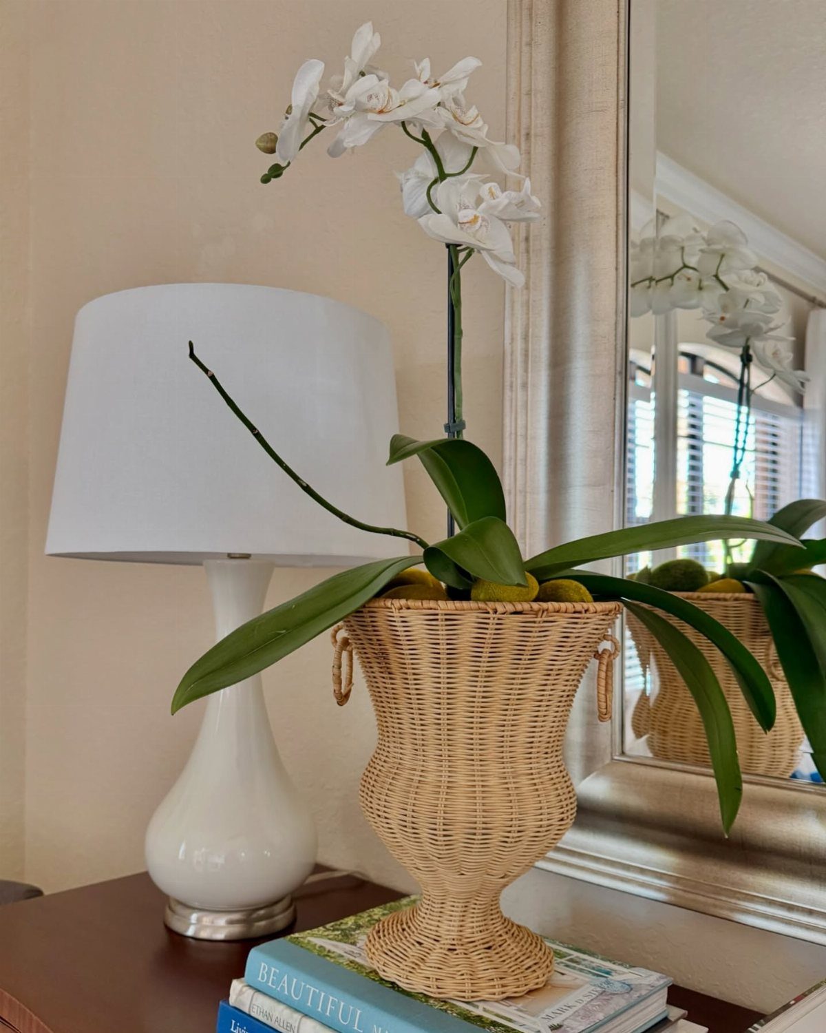 A light brown wicker urn vase holding white orchids sits next to a white lamp.