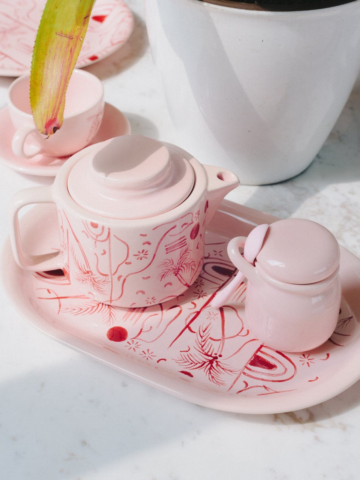 Pink ceramic teapot and sugar bowl with red tropical patterns on a matching tray.