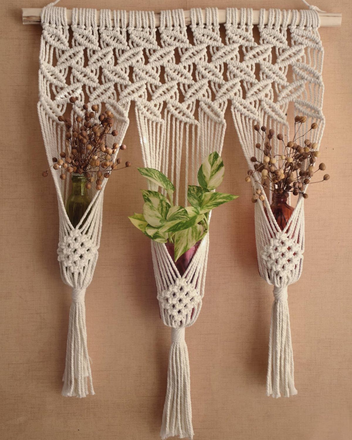 Three cream macrame planters hanging from a wooden rod containing greenery and dried flowers.