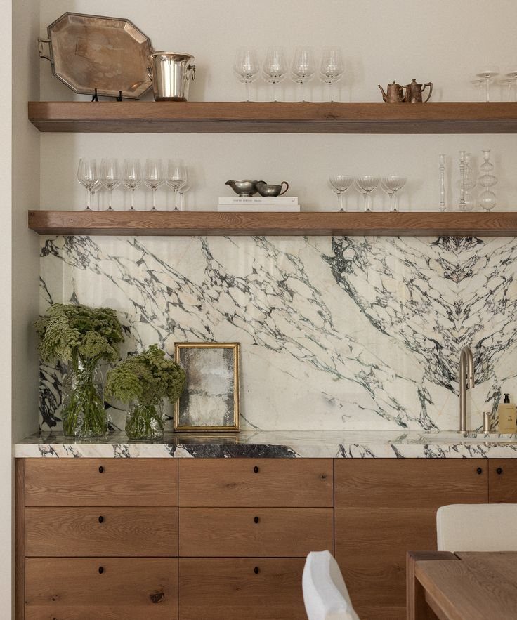 Wooden floating shelves holding glassware and silver decor above a marble countertop with green plants.