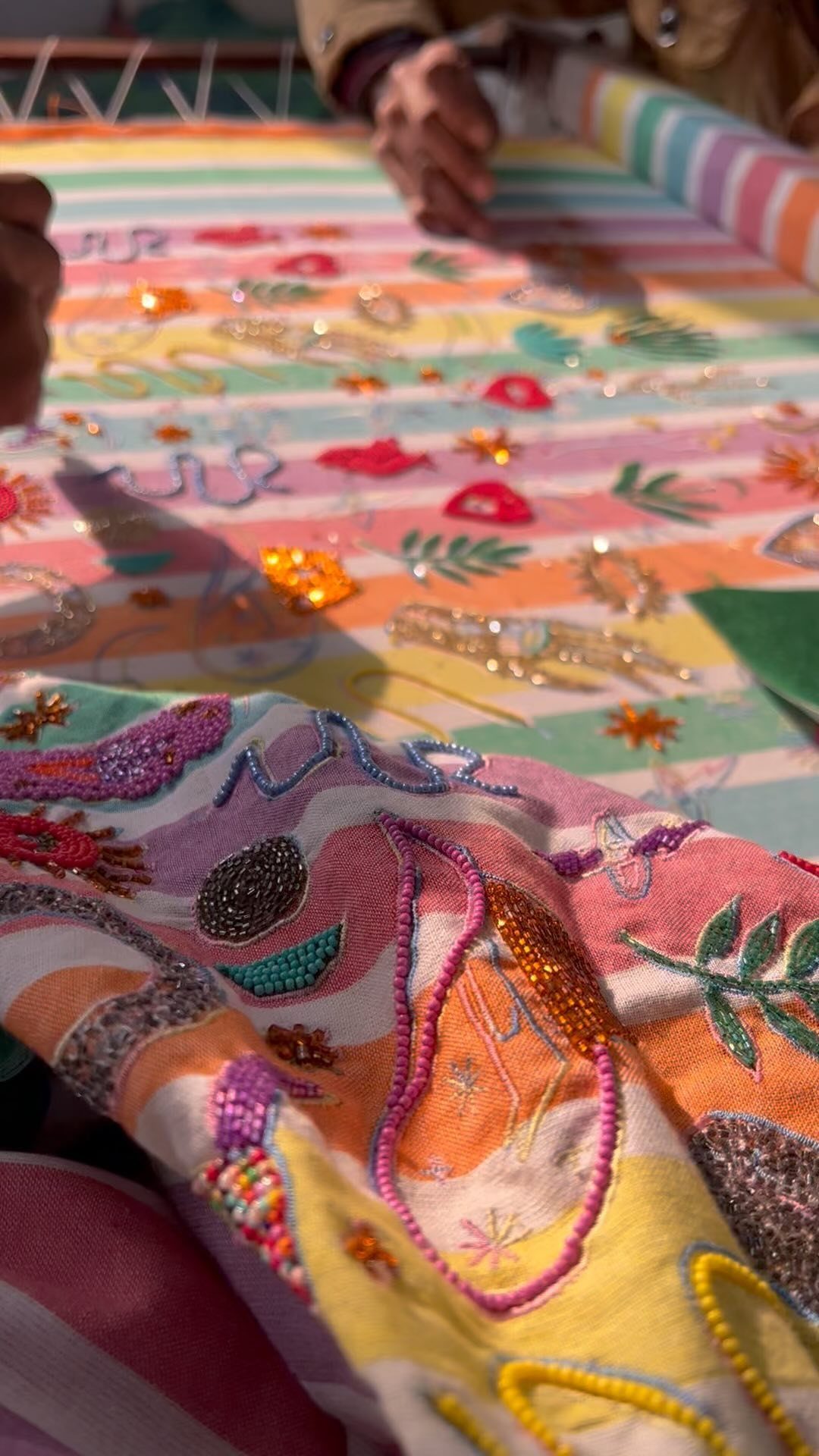 Hands decorating a vibrant, striped fabric with colorful beads and sequins in bright sunlight.