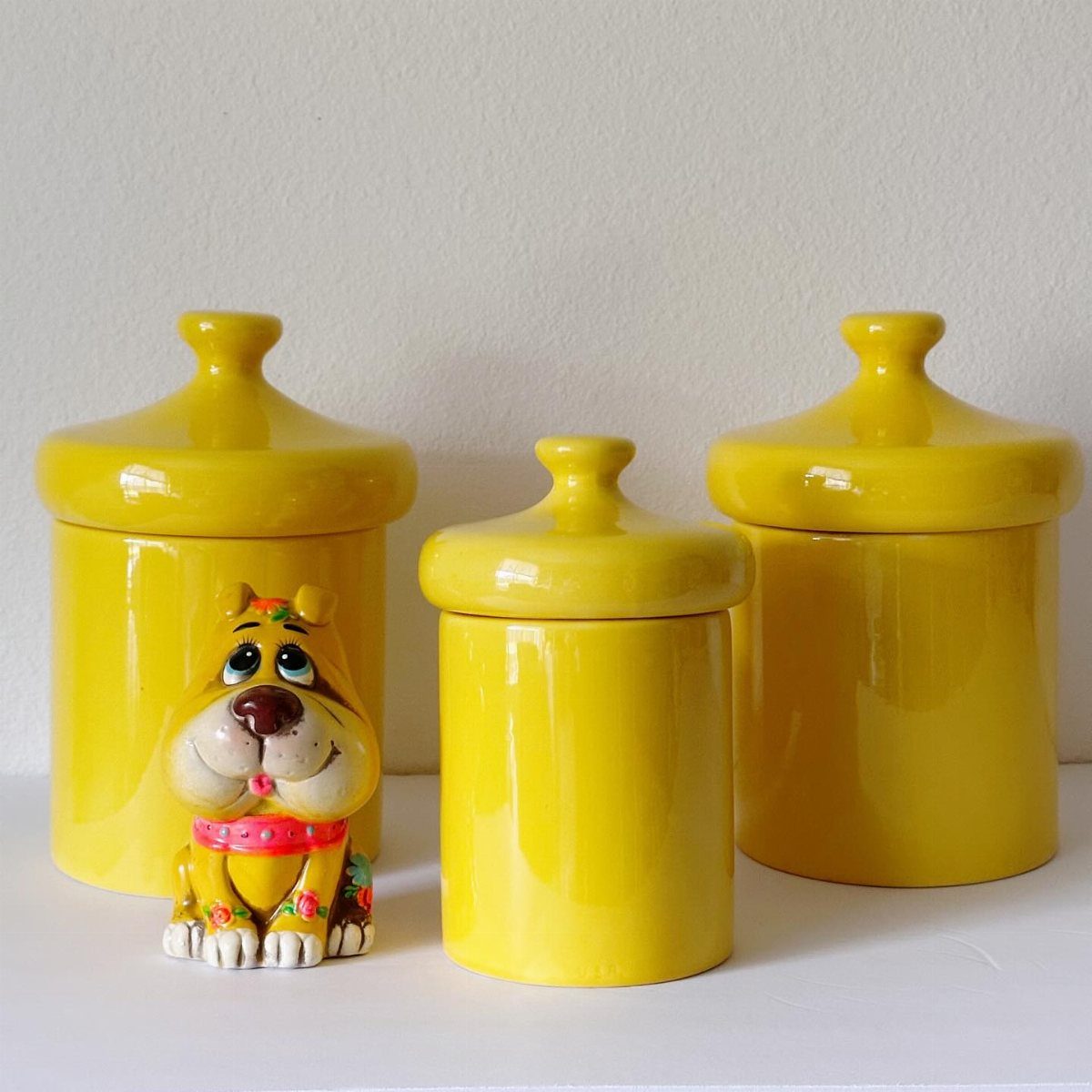 Three bright yellow ceramic canisters and a small colorful dog figurine on a white surface.