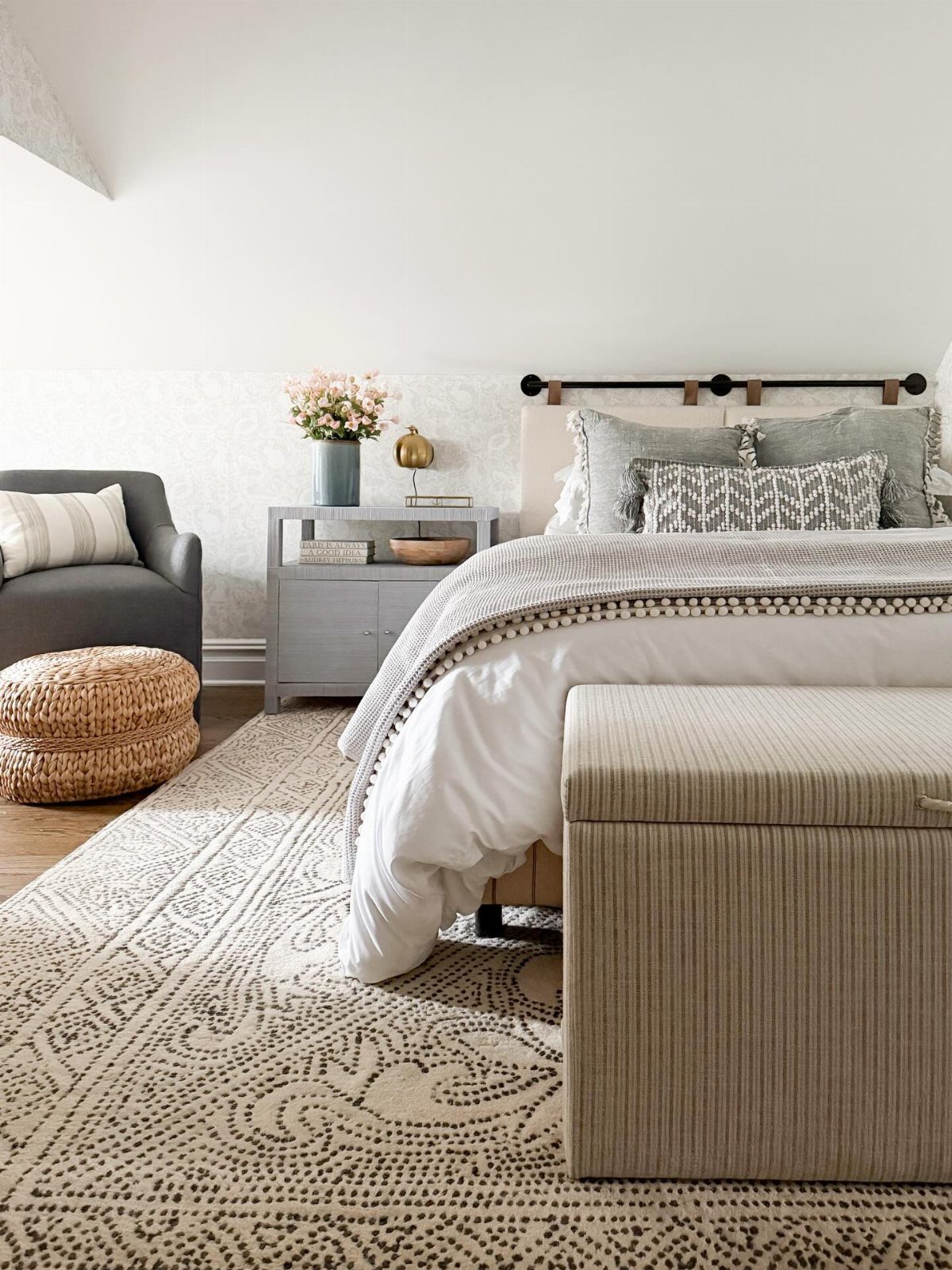 A cozy bedroom with white bedding, patterned pillows, a woven pouf, and a large patterned rug.