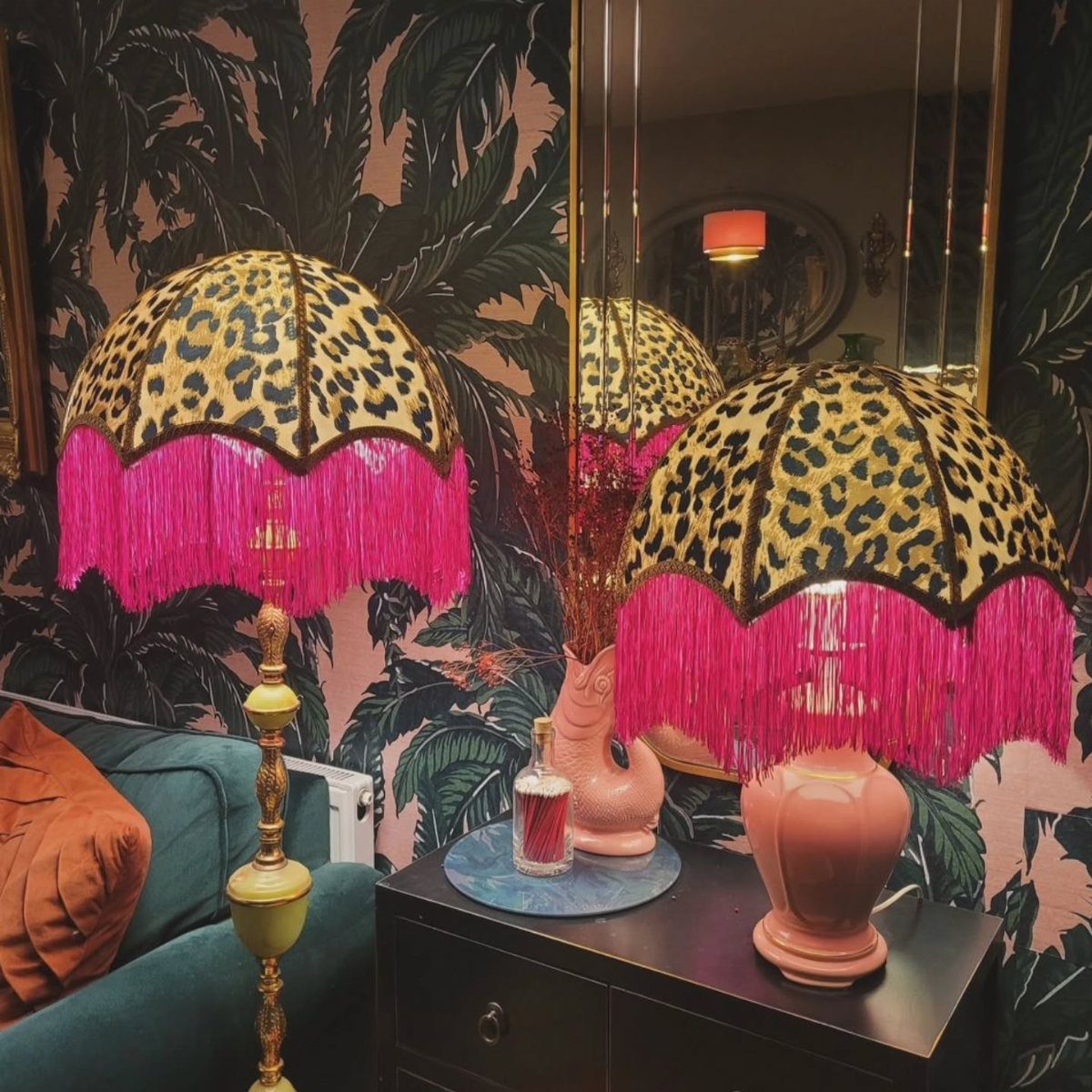 Leopard-print lampshades with pink fringe sitting on a dark wooden table against tropical wallpaper.