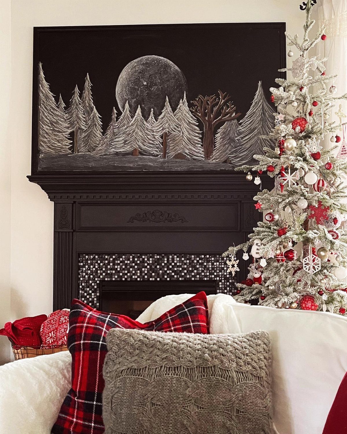 A snowy forest landscape painting hangs above a black mantel next to a decorated white Christmas tree.