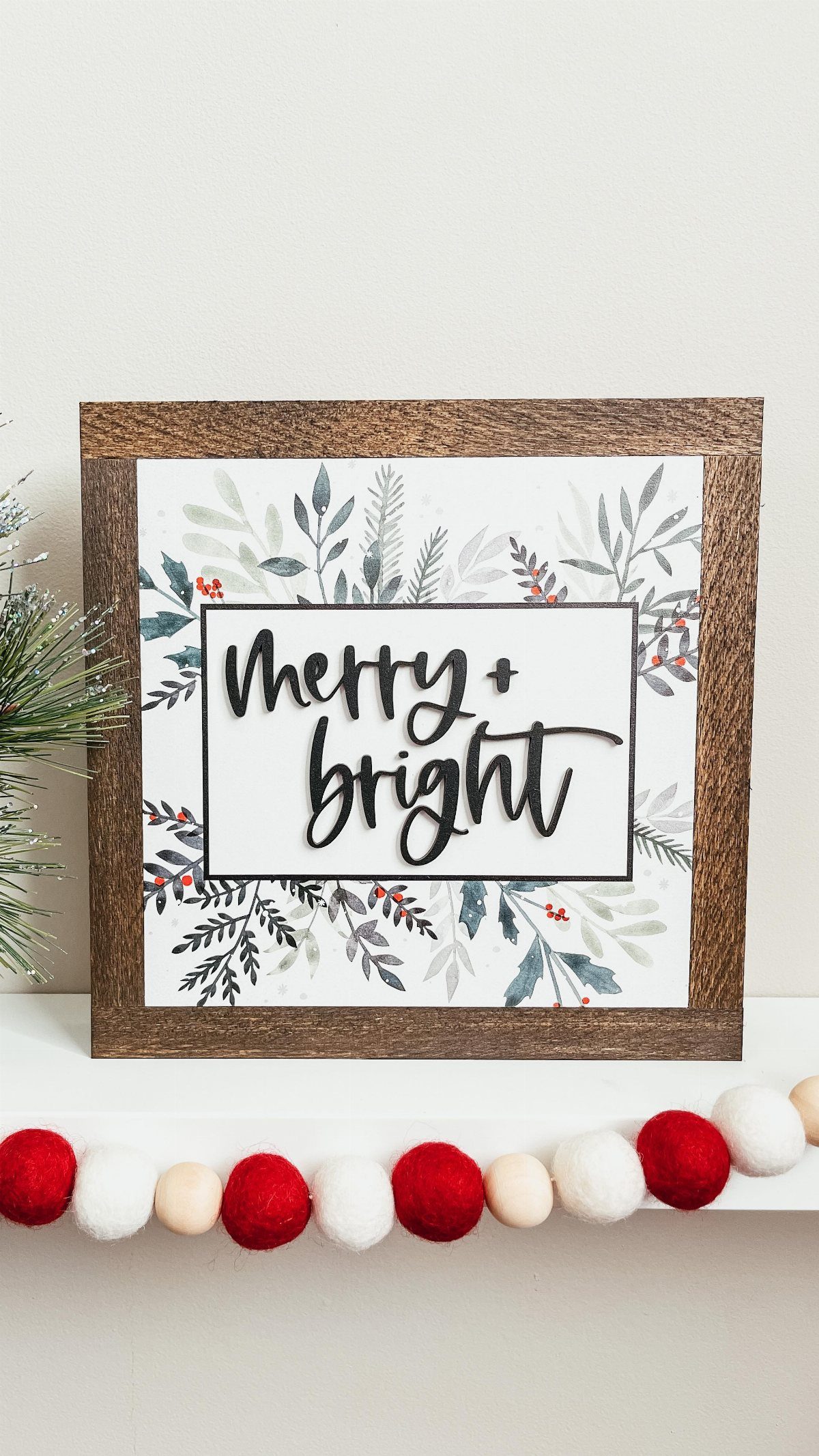 A wooden framed sign reading Merry + bright surrounded by botanical leaf illustrations.