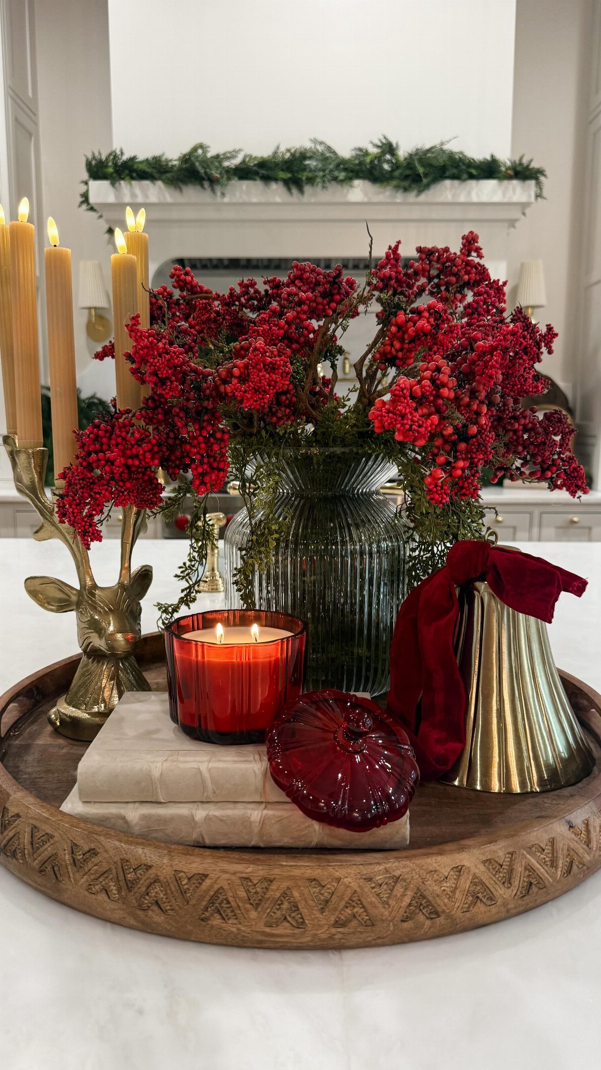 Red berries in a glass vase sit on a wooden tray with candles and gold accents.