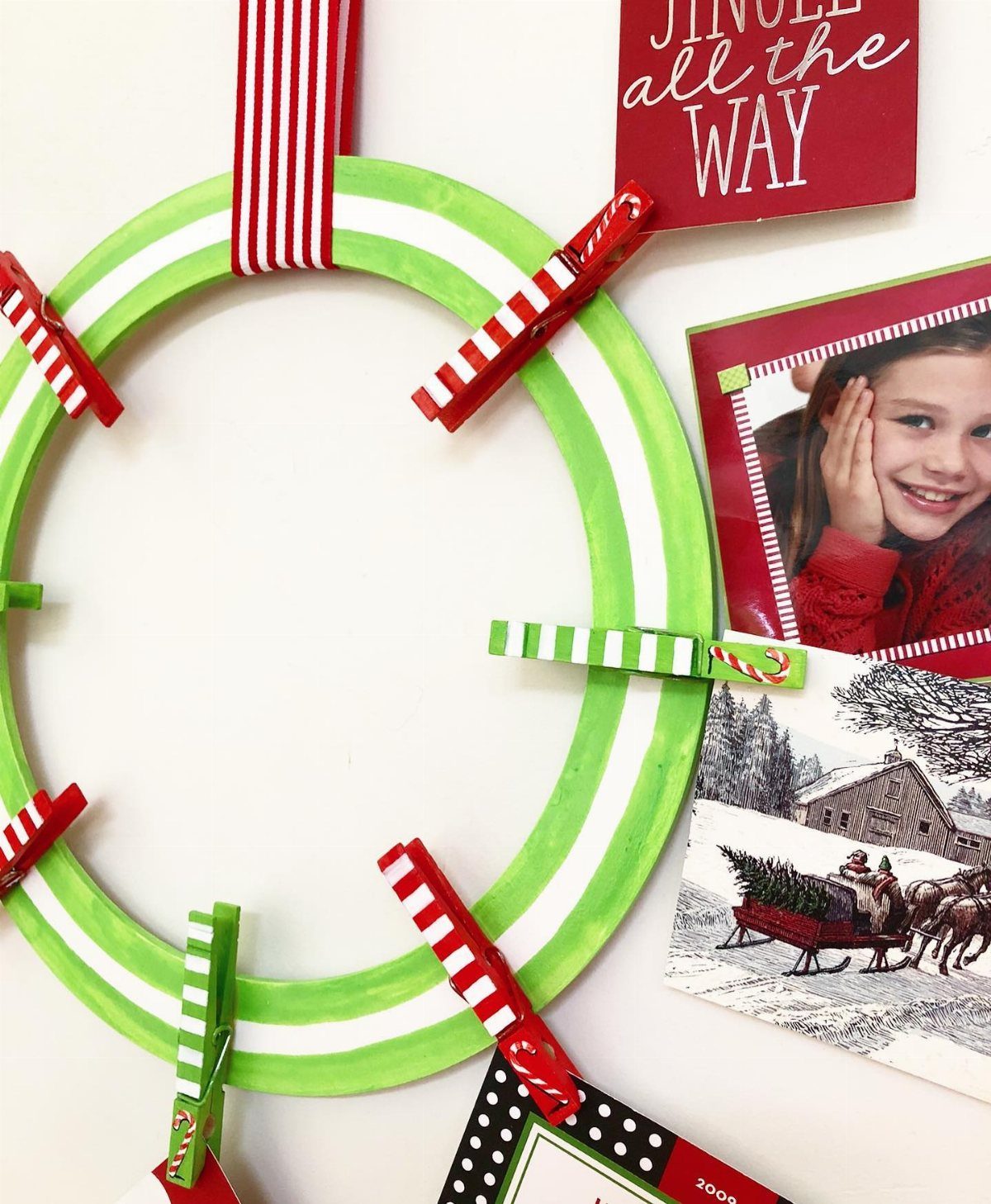 Green wreath with red and white ribbon holding various Christmas cards with clothespins.
