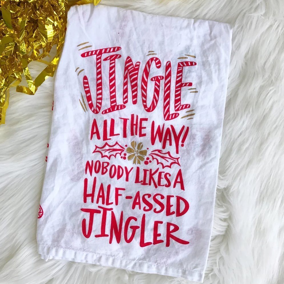 White kitchen towel with red holiday text resting on white faux fur with gold tinsel.
