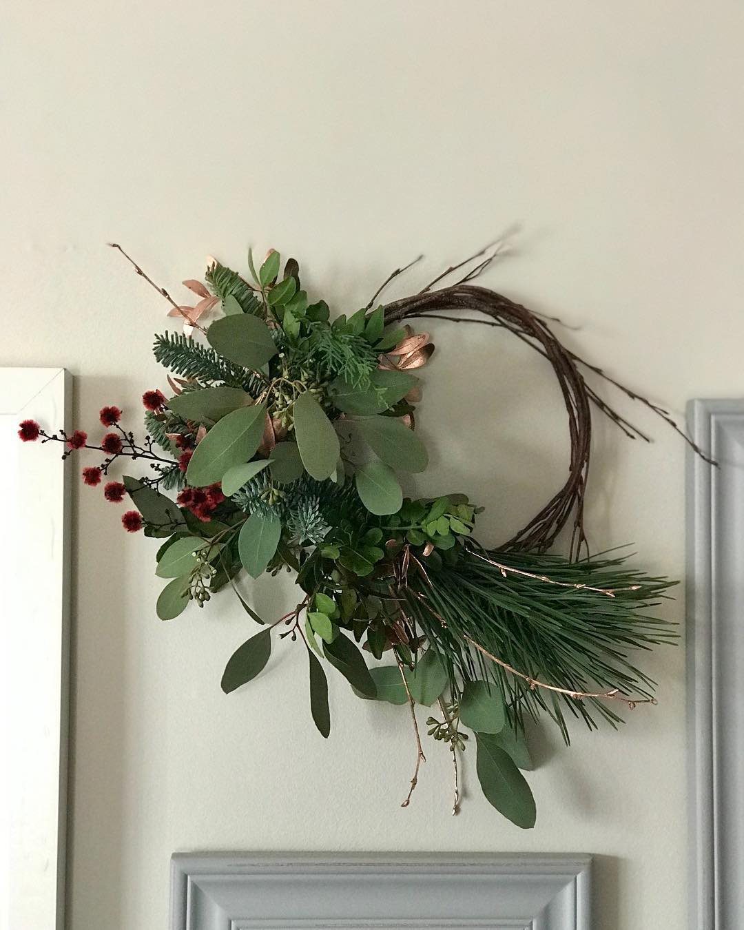 A grapevine wreath with eucalyptus, pine, and red berries hanging on a wall.