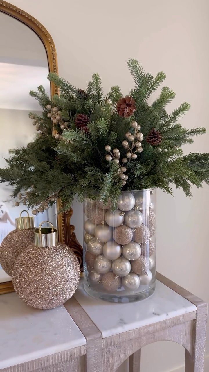 Evergreen branches with pinecones and gold berries in a vase filled with champagne ornaments.