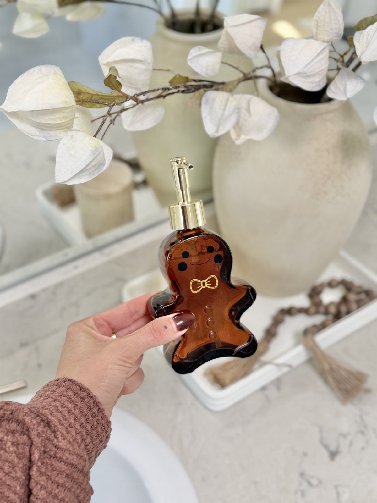 A hand holding a gingerbread man shaped amber glass soap dispenser against a marble background.
