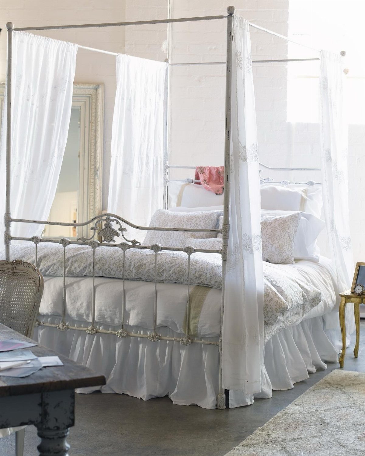 White metal canopy bed with sheer white curtains and light bedding in a bright room.