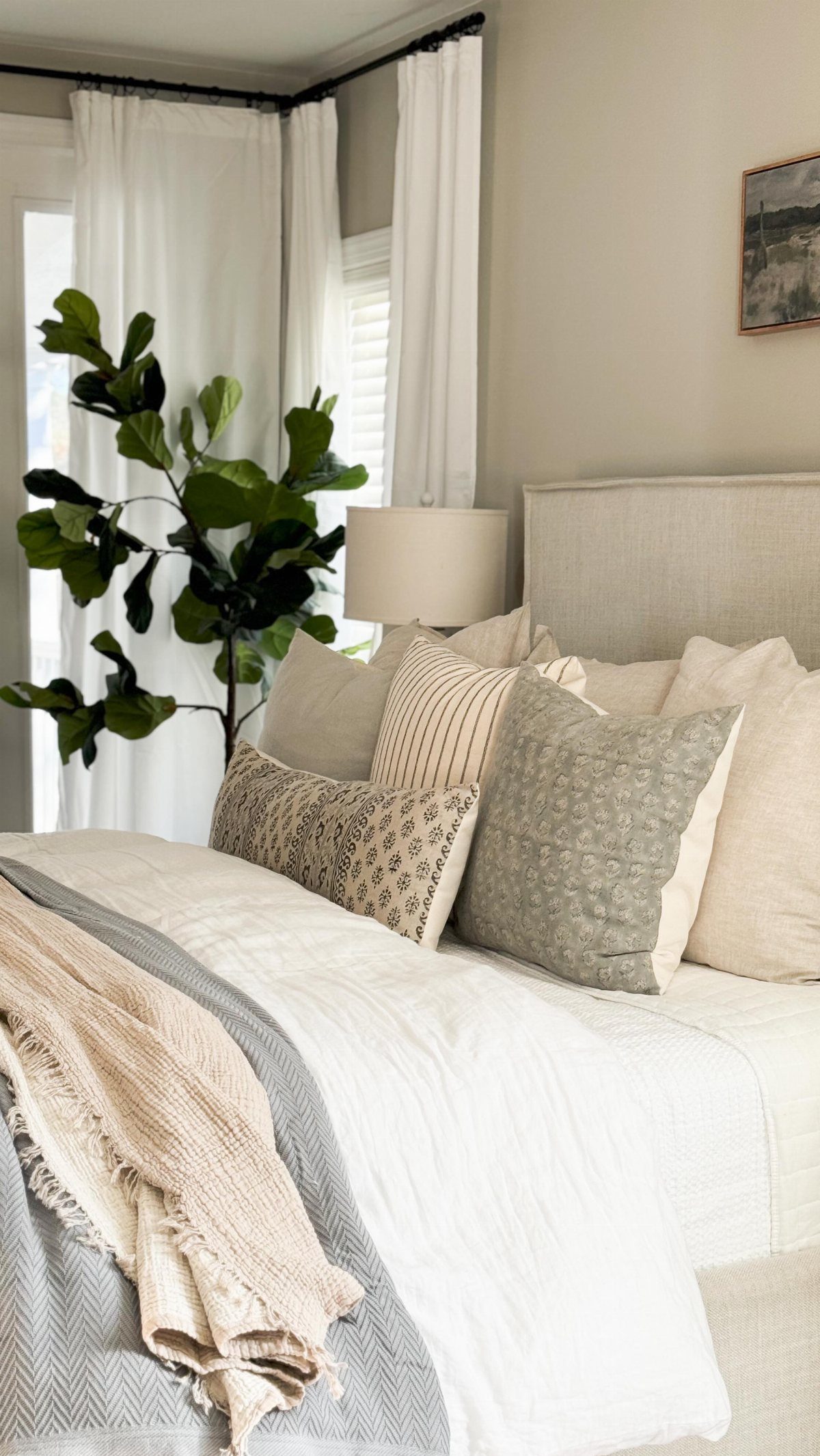 Layered neutral bedding with patterned pillows and a large green plant in a bright bedroom.