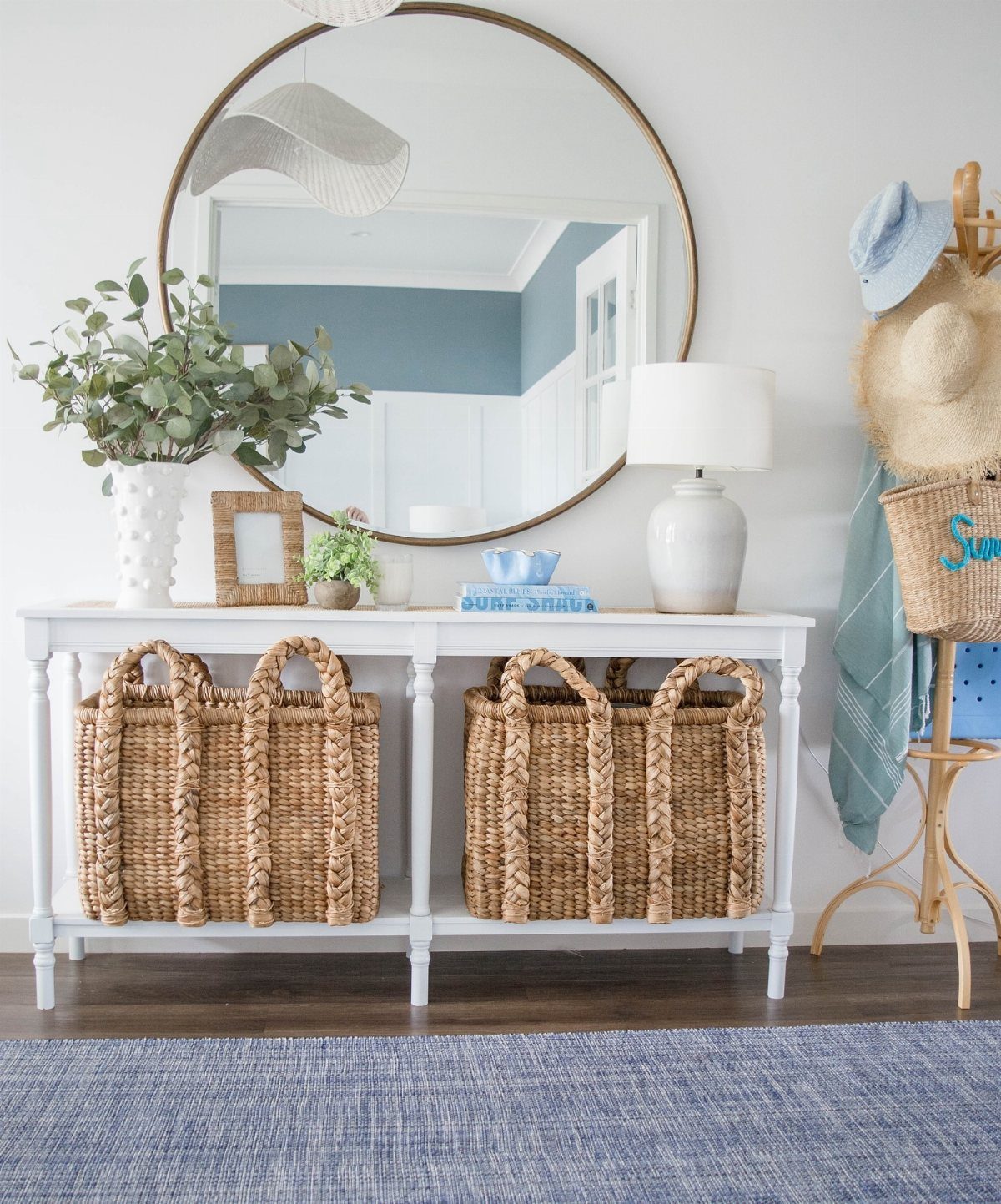 White console table with a round mirror, lamp, and large woven baskets in a coastal entryway.