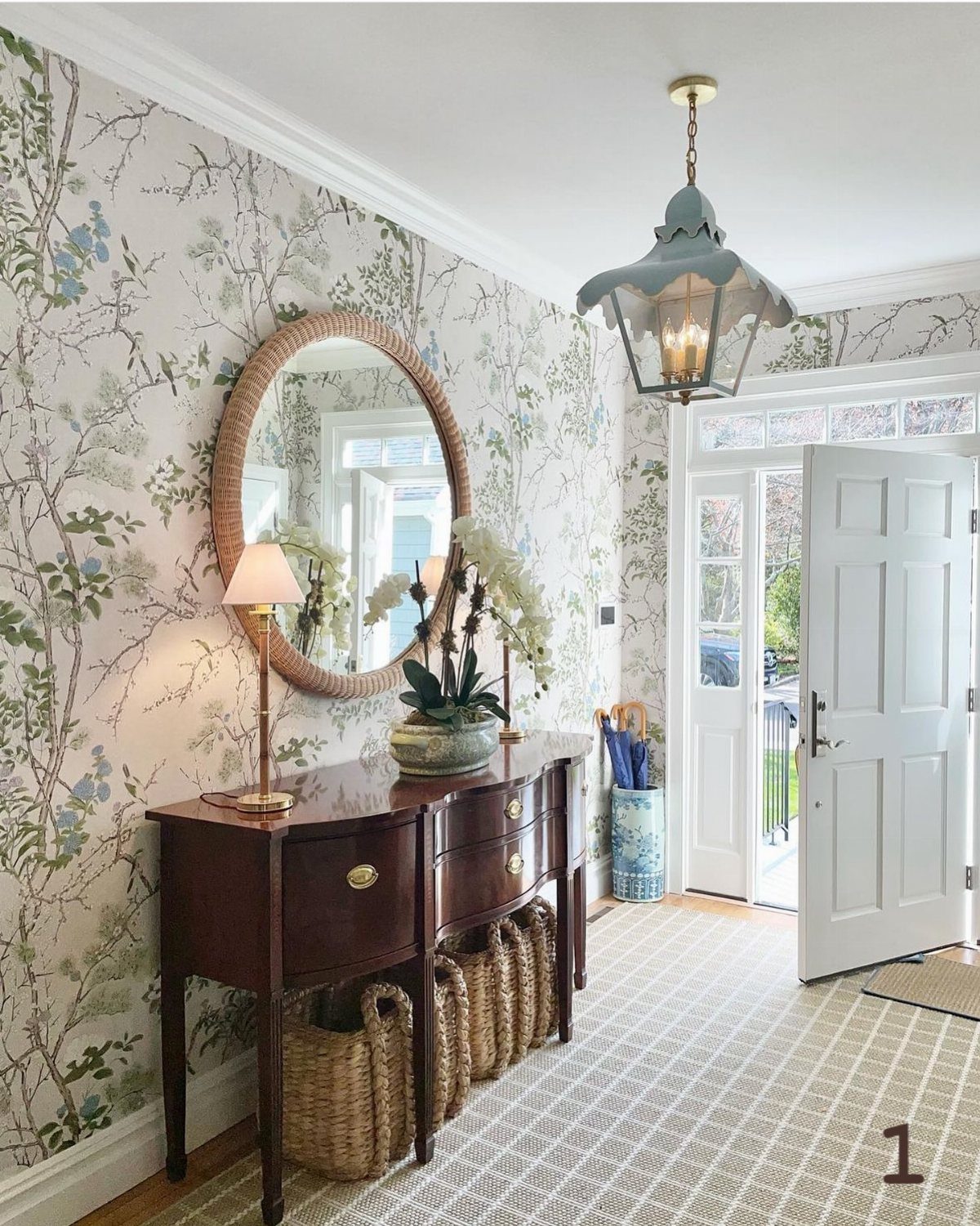 A dark wood console table with a round woven mirror against floral wallpaper in a bright entryway.