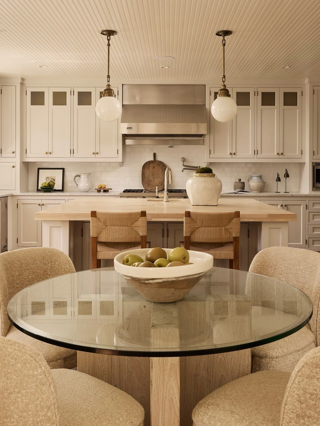 Glass dining table with fruit bowl in a bright, coastal-style kitchen with rattan seating.