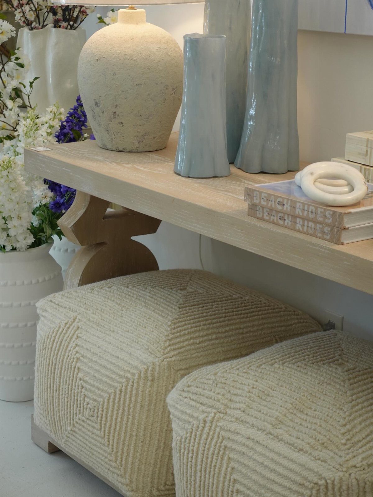 Light wood console table with cream ceramic vases and two textured fabric poufs underneath.