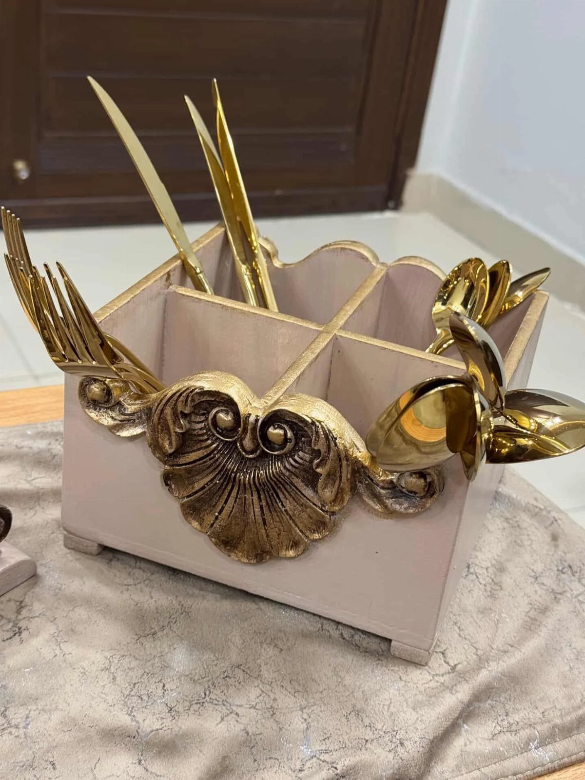 Ornate beige and gold sculptural organizer holding long gold utensils on a textured surface.