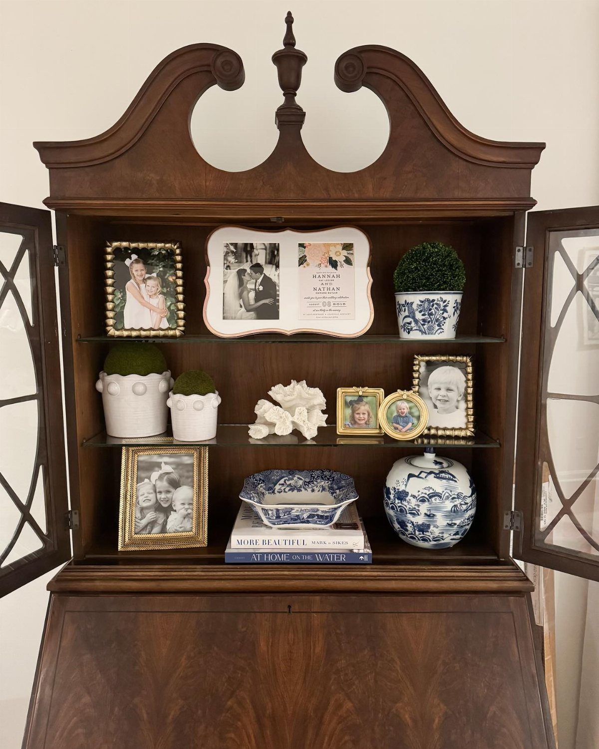 Vintage wooden secretary desk displaying gold frames, coral, and blue and white ceramics on glass shelves.
