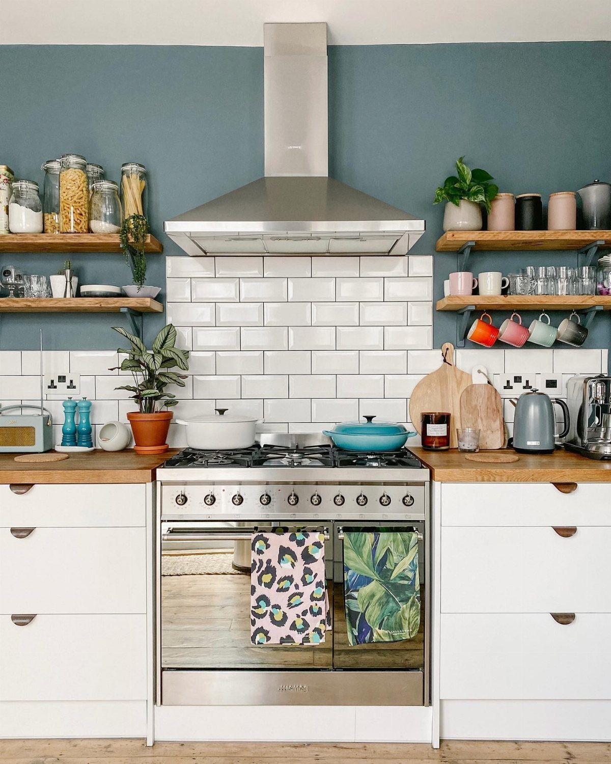Wooden open shelves holding jars, plants, and mugs against a blue wall and white tile backsplash.