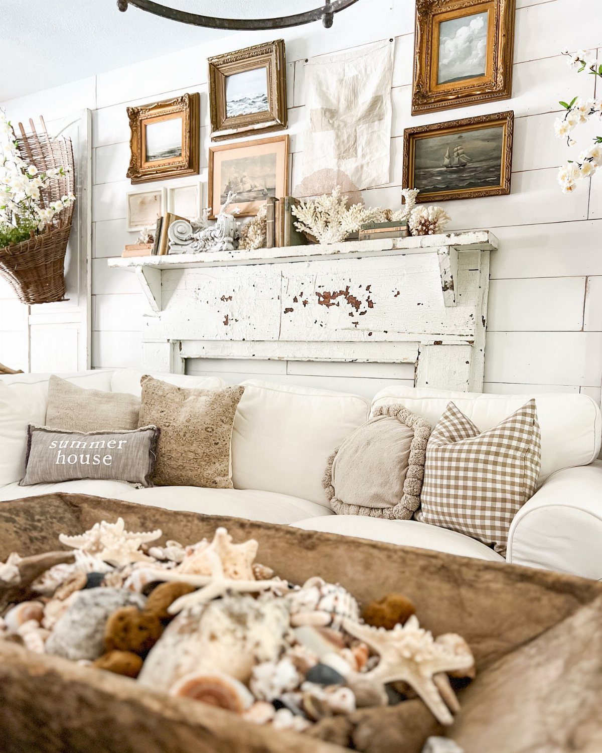 Distressed white mantelpiece with vintage gold frames and seashells above a white sofa.