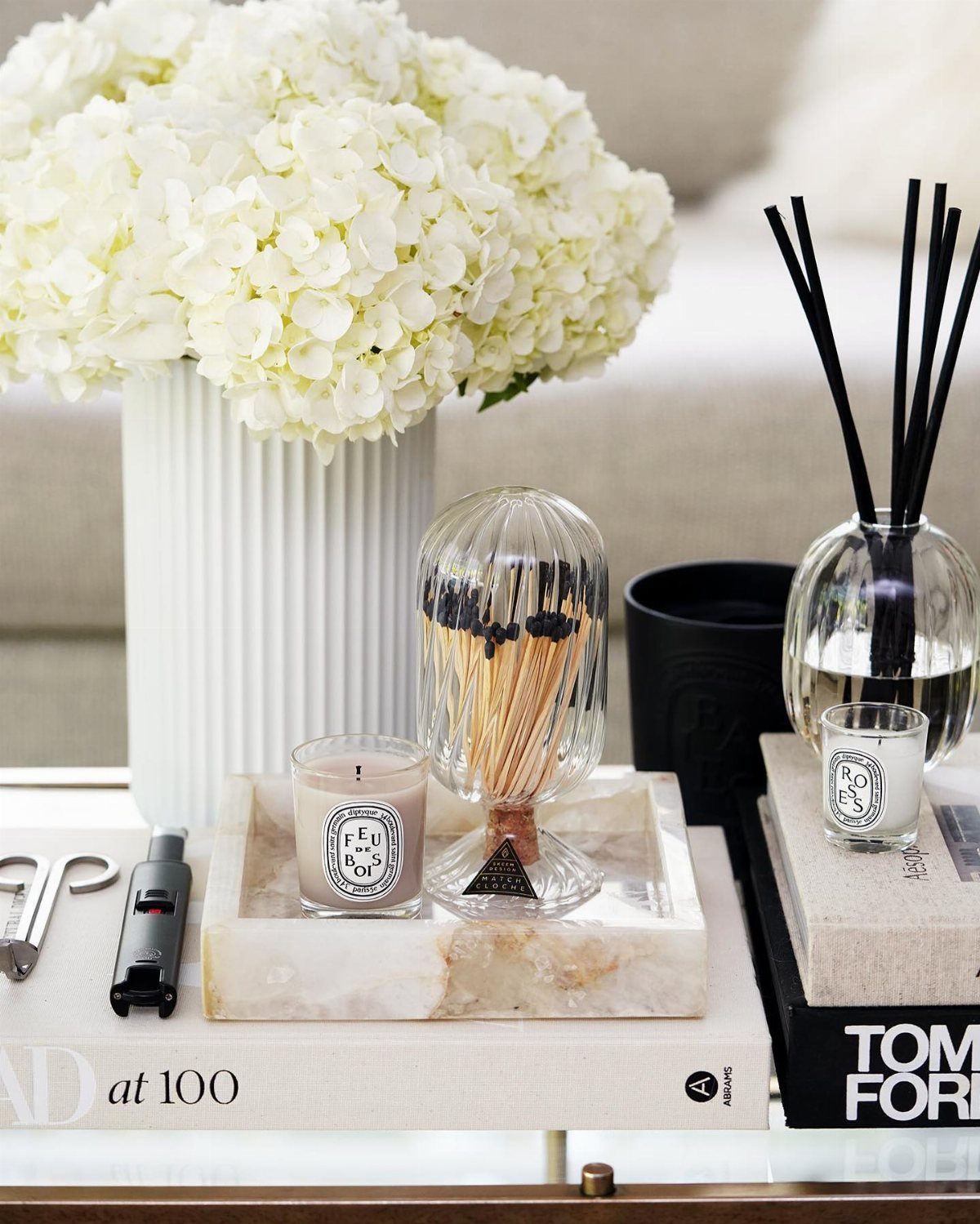 White hydrangeas in a fluted vase next to a glass match cloche on a marble tray.