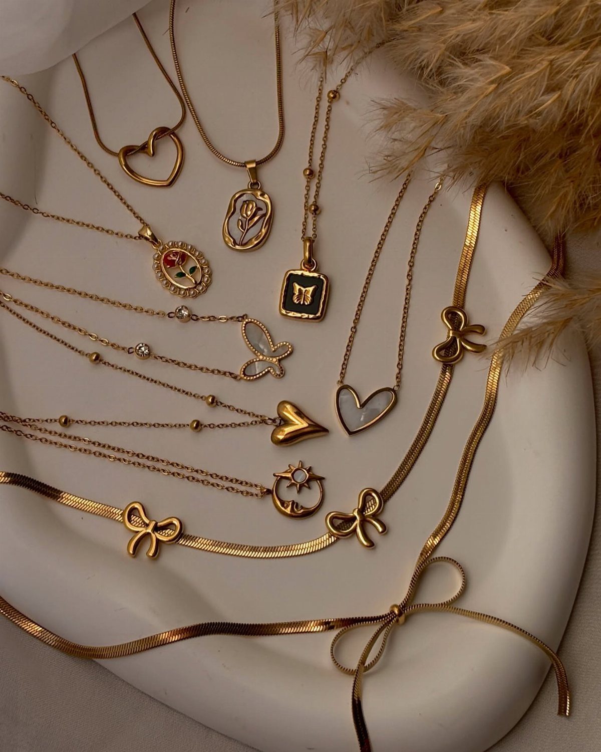 Gold necklaces with various pendants arranged on a white ceramic tray next to dried grass.