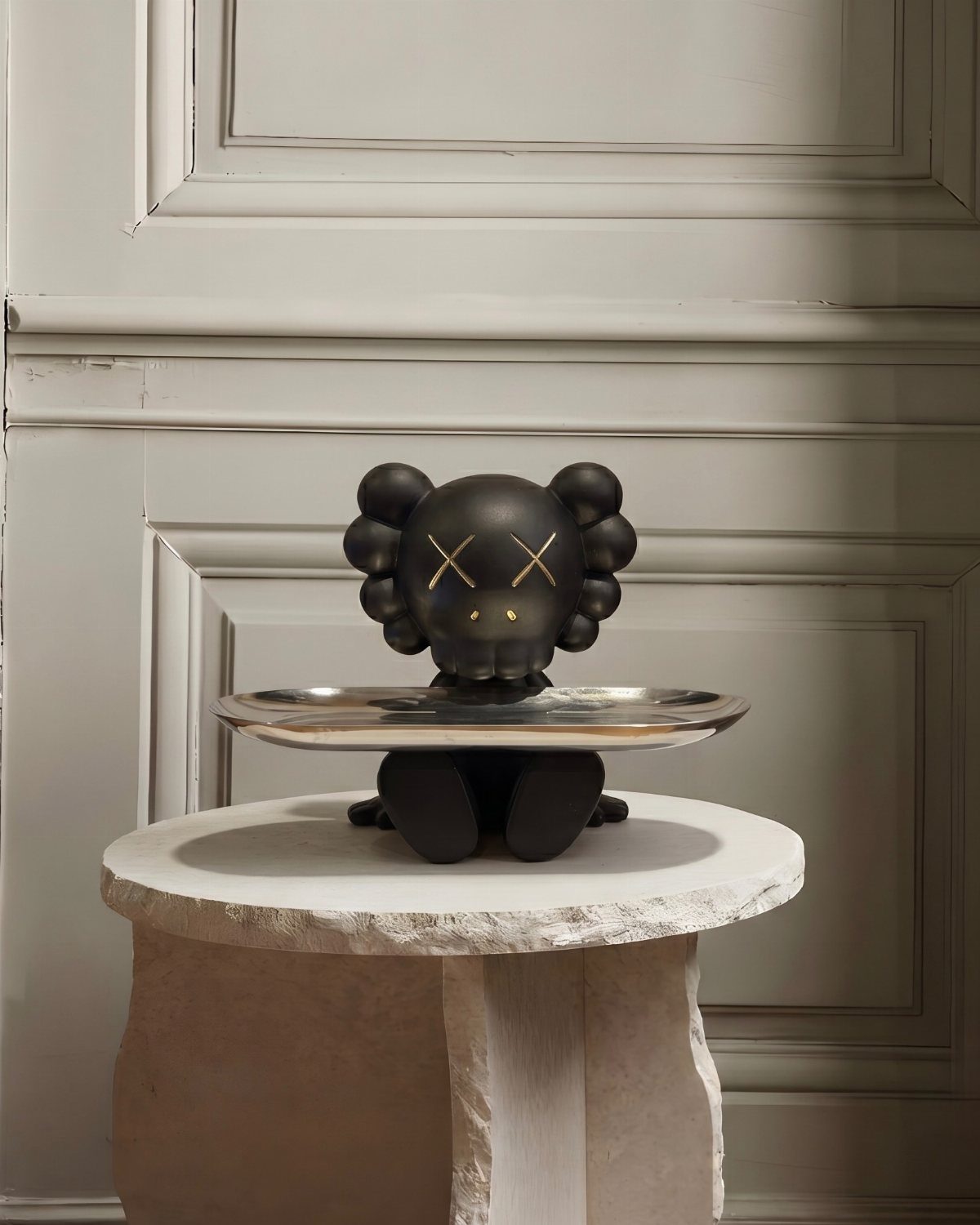 Black character sculpture with a silver tray resting on a white stone pedestal against paneled walls.
