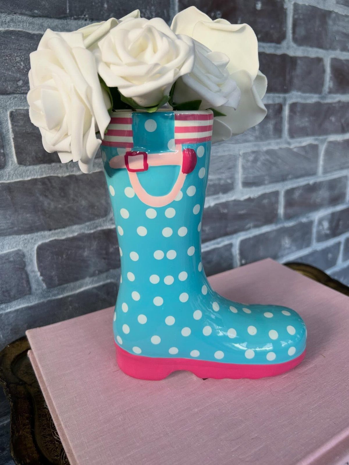 A blue polka-dot rain boot vase filled with white roses on a pink surface.