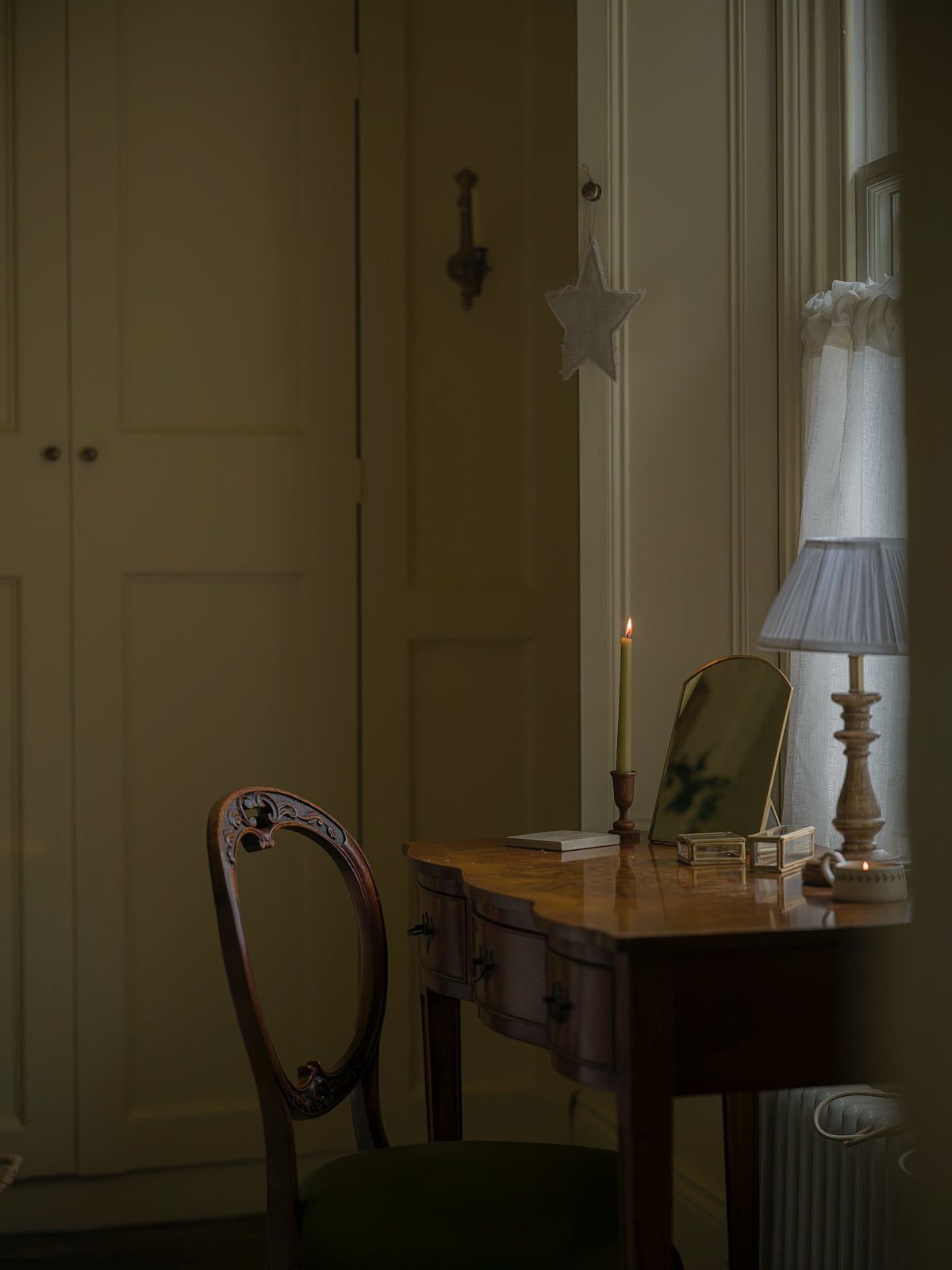 A wooden desk with a lit candle, lamp, and mirror sits in a cozy, dimly lit room.