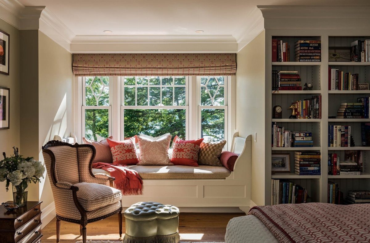 A cozy window seat with colorful pillows and an armchair next to a large bookshelf.