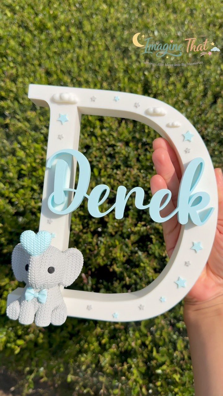 A hand holding a large white decorative letter D with the name Derek and a small crochet elephant.