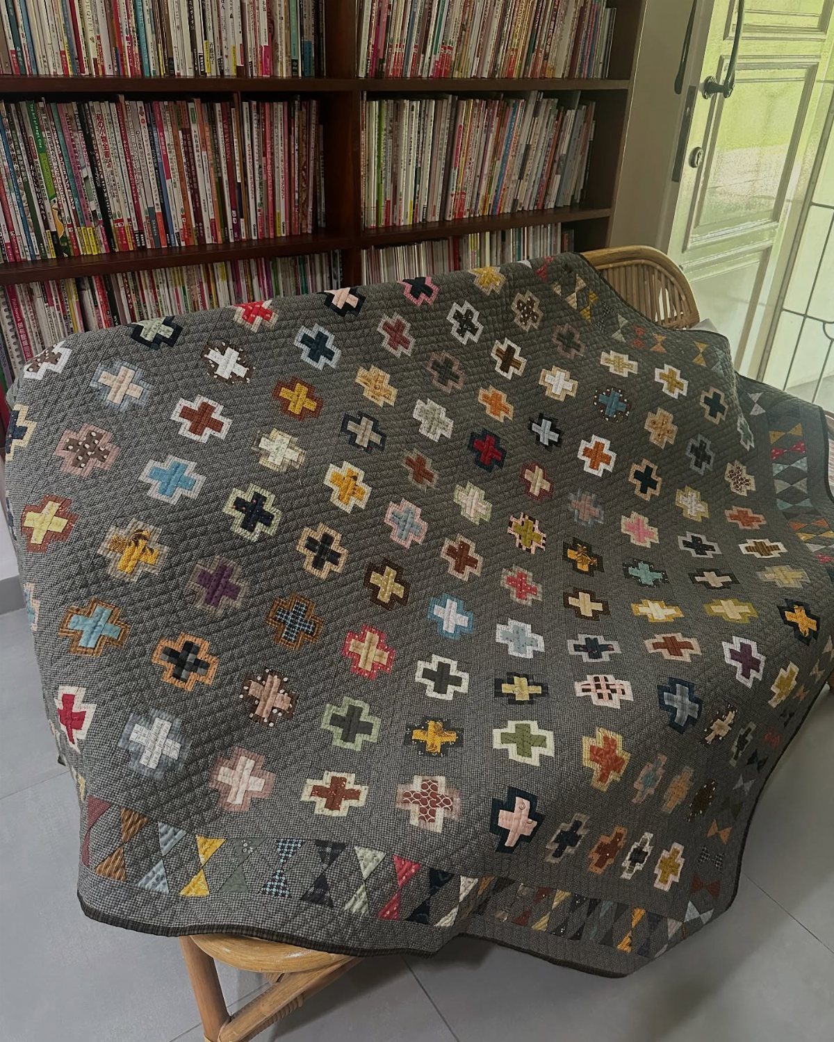 A grey quilt with colorful cross patterns draped over a wooden chair near a bookshelf.