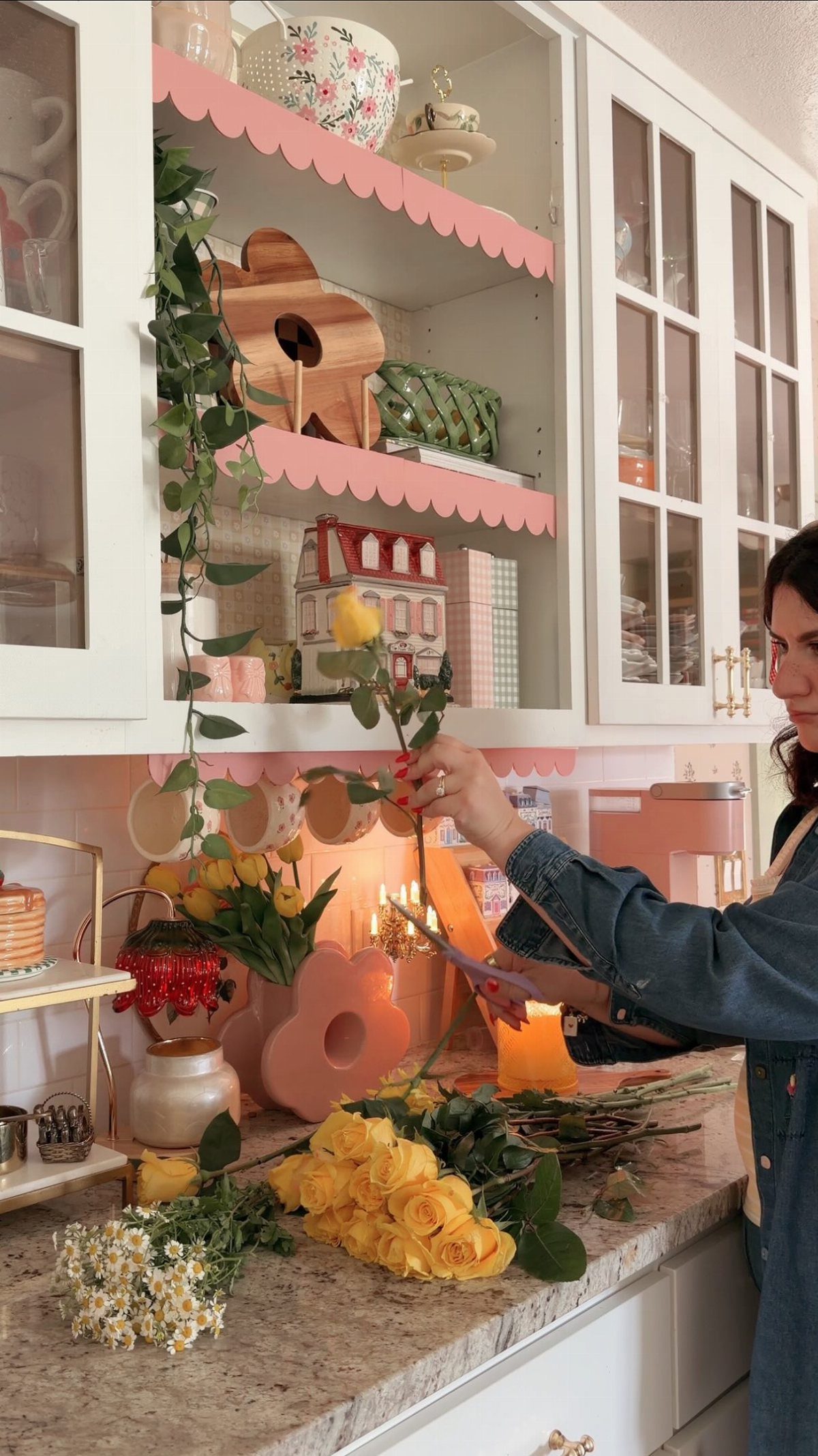 A person holding a yellow rose in a kitchen with pink scalloped shelving and white cabinets.