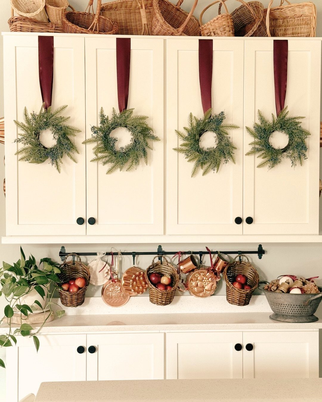 Four small evergreen wreaths with burgundy ribbons hanging on cream kitchen cabinets above fruit baskets.