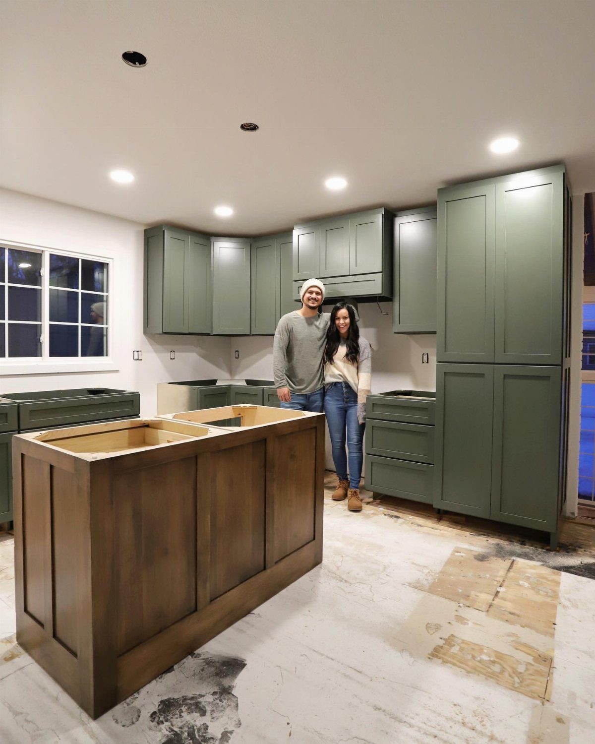 A couple standing in a kitchen featuring sage-green cabinets and a dark wood island.