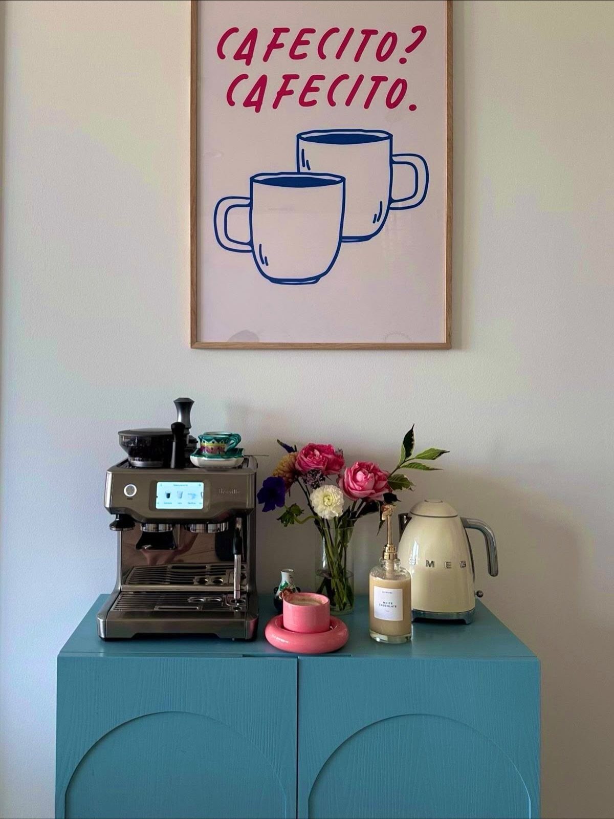 Espresso machine, flowers, and a kettle arranged on a blue sideboard under framed art.