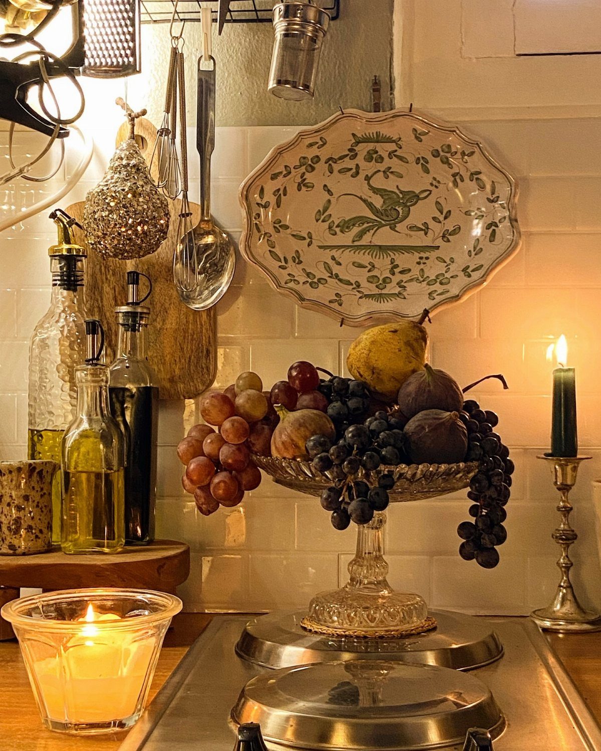 Fruit in a glass bowl sits near a decorative ceramic plate and lit candles.