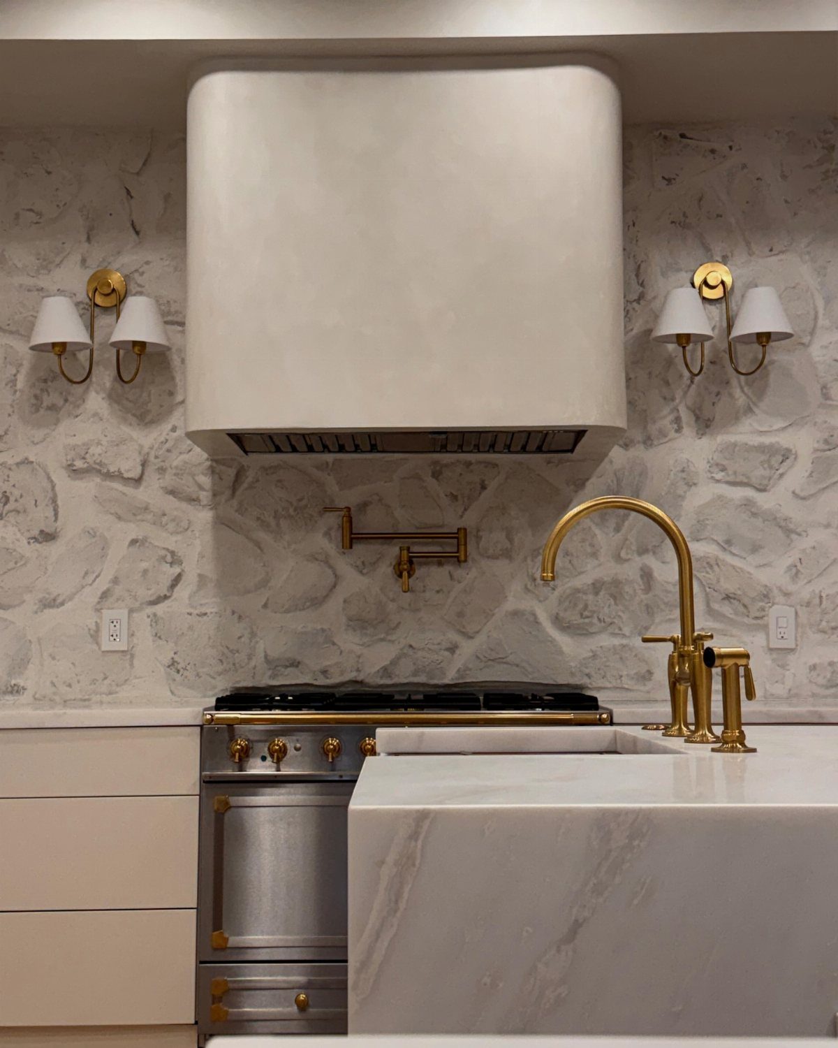 Stone textured backsplash behind a large plaster range hood and gold kitchen fixtures.