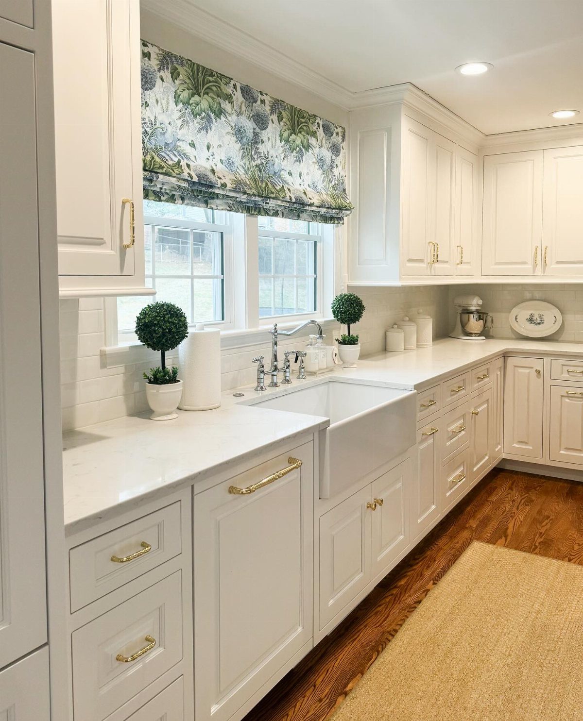 White kitchen cabinets and marble countertops with a floral fabric Roman shade over the window.