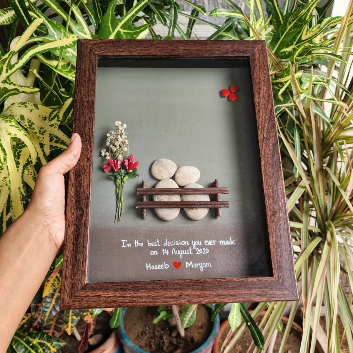 A hand holding a framed piece of personalized stone and flower art against green plants.