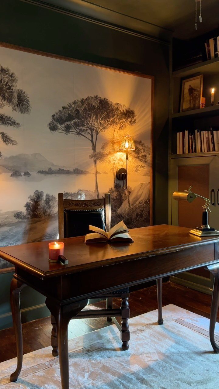 A vintage wooden desk with an open book and candle against a landscape mural.