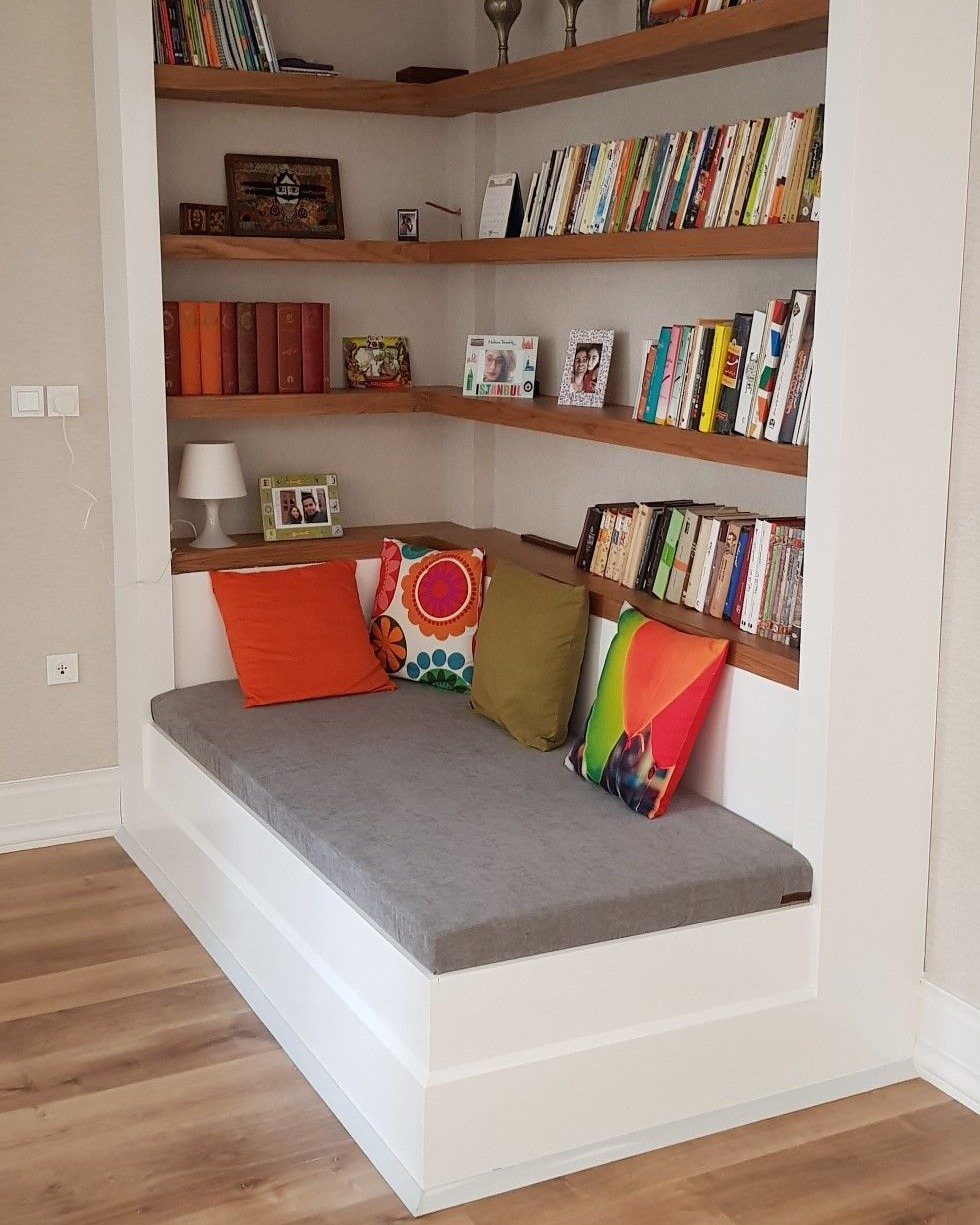 Built-in corner reading nook with grey cushioned bench, colorful pillows, and wooden bookshelves filled with books.